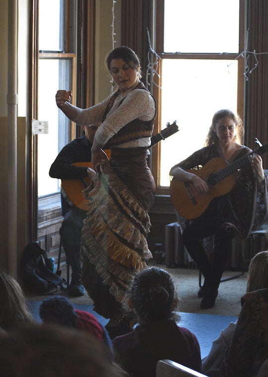 A woman holds her long ruffled skirt as she dances in front of a room of people.