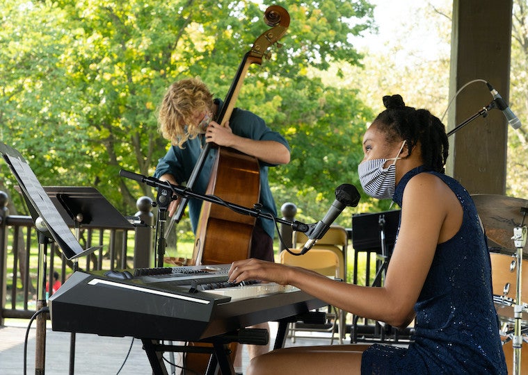 A female student plays the piano and sings into a microphone while a male student plays the cello next to her.