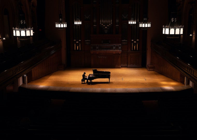 A male student plays piano on a large concert stage.