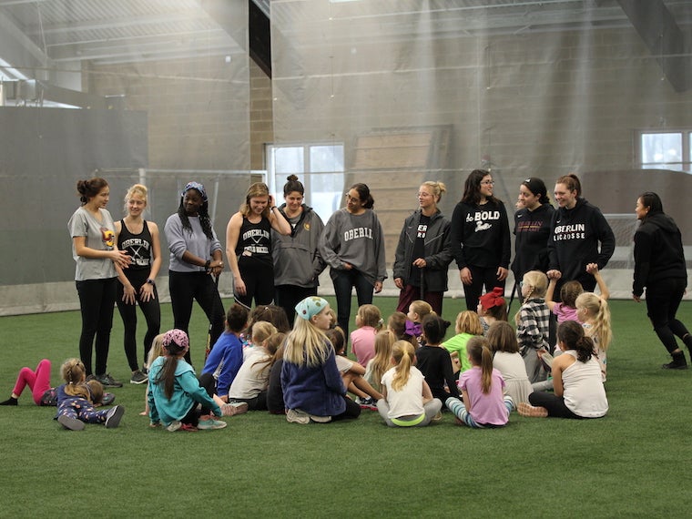 A group of children sit on the floor in an athletic facility with college athletes standing in front of them.