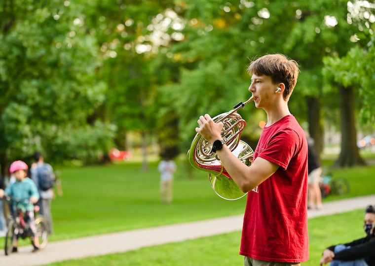 A student plays a French horn in a park.