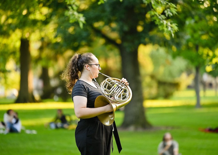 A student plays a French horn in a park.