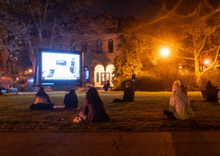 Students sit on a lawn at night and look at a large screen.