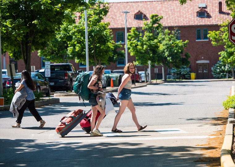 Three students with suitcases walk across a parking lot.