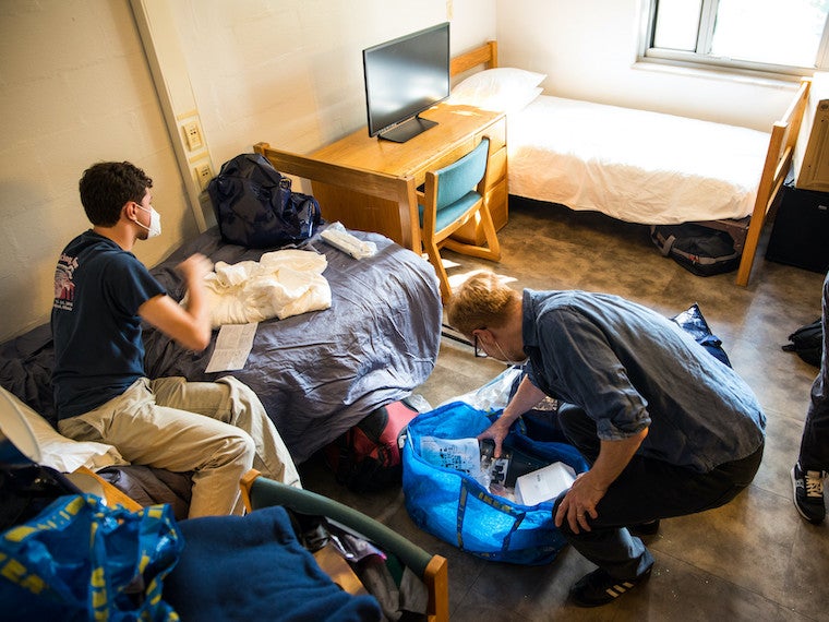 A man and college student unpack luggage in a dorm room.
