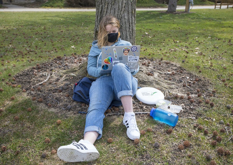 A girl leans against a tree and types on a laptop.