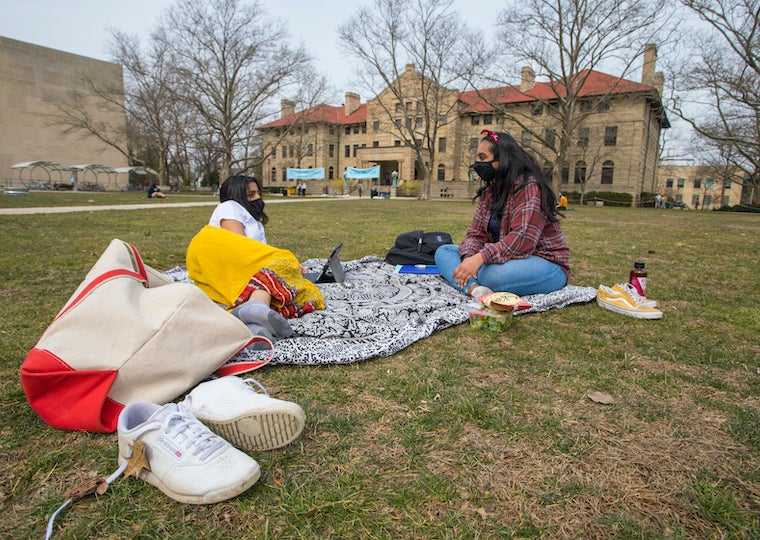 Two students share a tapestry blanket and relax on a lawn.