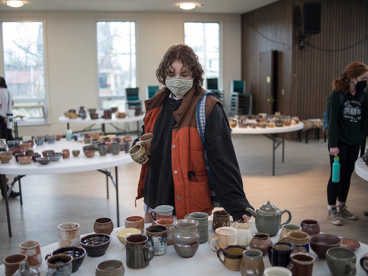 A college student looks at a table with lots of mugs on it.