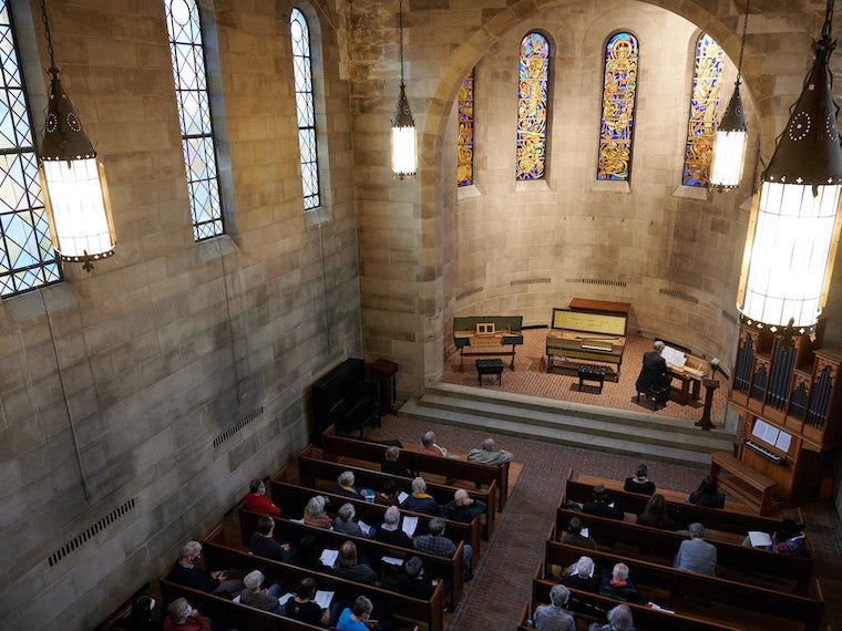 A small audience is seated in a stone chapel.