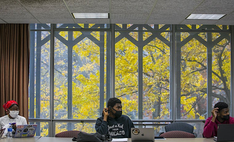 Three students sit behind large windows with trees.