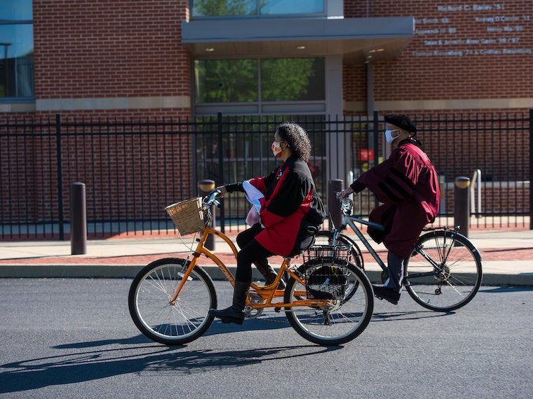 Two professors in graduations gowns ride bikes.