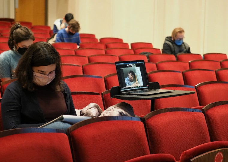 A masked student sits next to a laptop monitor that shows a student on the screen.