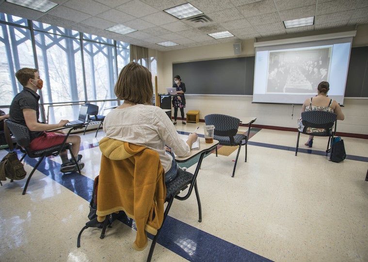 Students sit at desk in a classroom and face a projector screen with images.