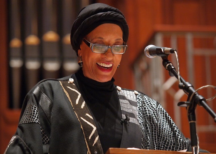 A woman wearing a head wrap speaks bows her head in front of a podium.