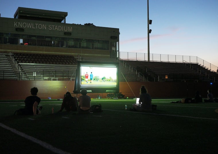 Student sit on a football field and watch a movie on a big screen. 