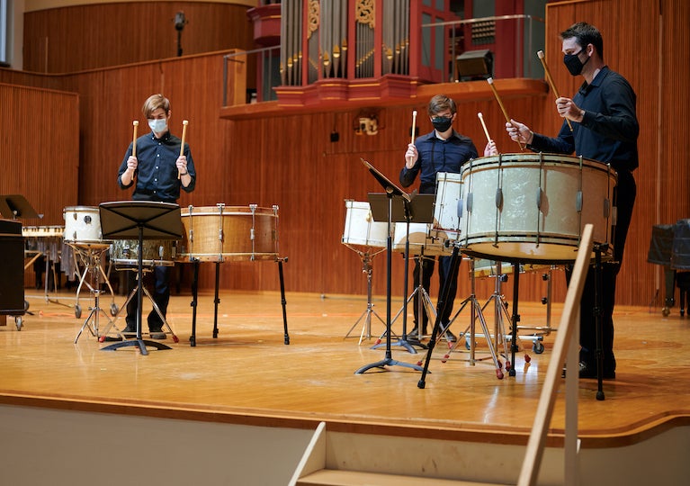 Three students play kettle drums on a stage.