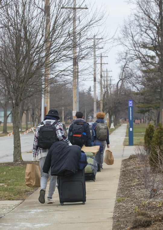 A line of students walk down a sidewalk with luggage.