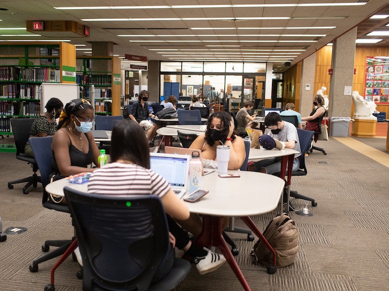 Groups of students study at tables in a library.