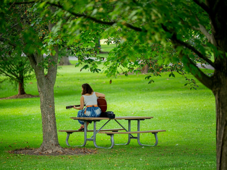 A girl plays a guitar in a park while skiing on a picnic table.