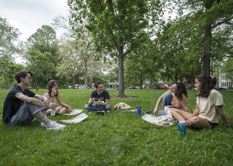 A group of students sit in a semicircle while eating lunch in a park.