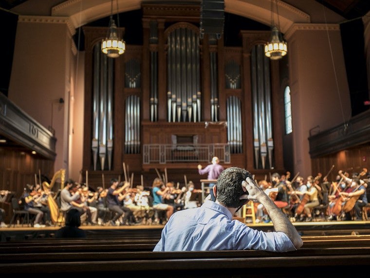 A man sits in a large auditorium by himself while looking at a large orchestra on stage.