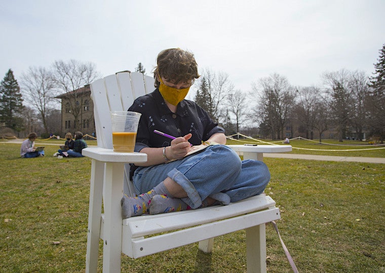 A student writes in a notebook while sitting in a large wooden lawn chair.