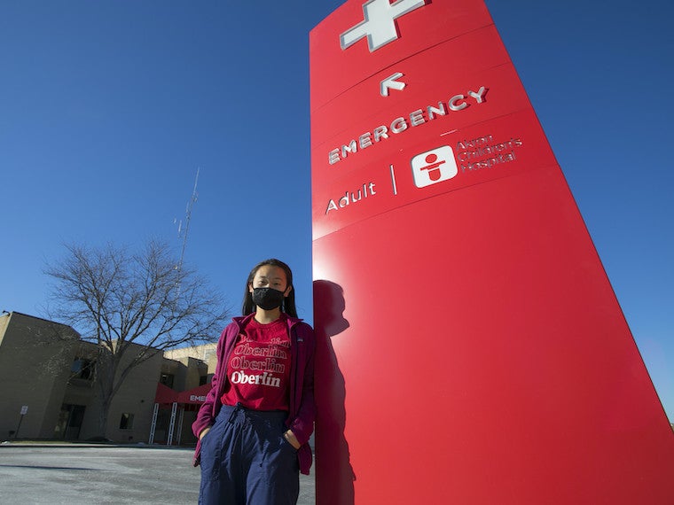 A girl leans against an emergency room sign in front of a hospital.