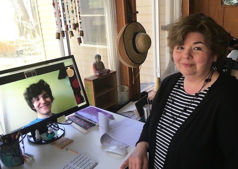 A woman stands next to a laptop that has a picture of a student on the screne.