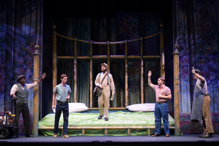 A large bed on a stage with five men standing on and near it.