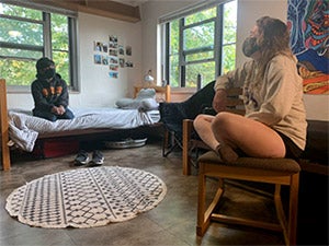 Two friends have a conversation in a dorm room, situated well apart from each other and wearing masks.