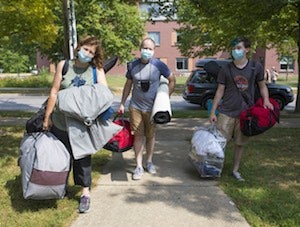 A student and his parents carry luggage. All are wearing masks.