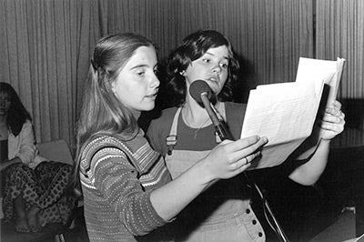 Two women speak into a microphone while holding a script. Black & white photo.