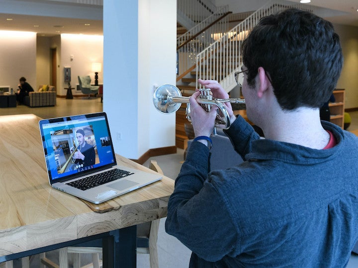 A trumpeter plays in front of his laptop, while another is on screen listening. 