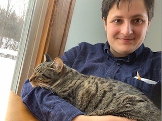 Benny (white masc person with brown, short hair) facing camera selfie-style with a sleeping tabby cat named Bruce in his arms.