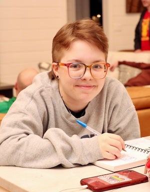 A picture of Cecil, sitting at a counter and drawing in a notebook. He is looking up at the camera and smiling. He is wearing a grey sweatshirt, has short brown hair, and rainbow glasses. 