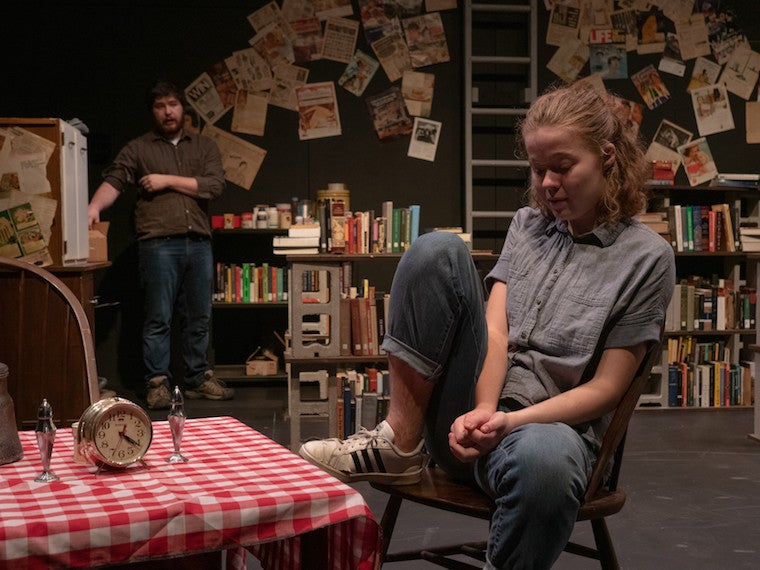 A girl sits in a chair with her head down and a man looks at her from the other side of the room.