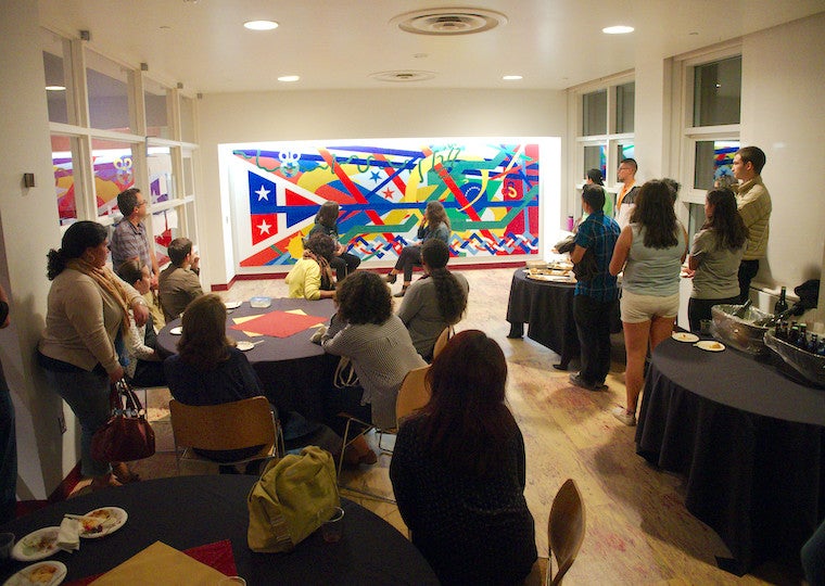Groups of people sit and tables and stand in front of a large mural.