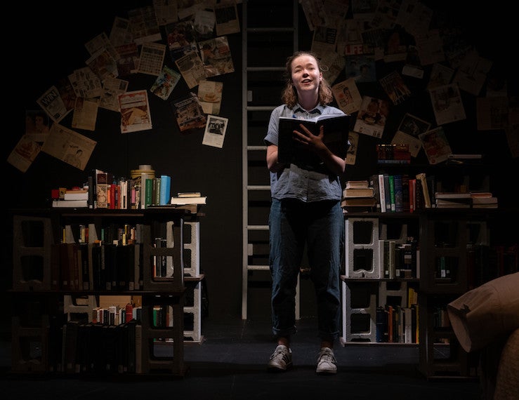 A girl smiles as she looks up into the spotlight while holding a notebook.
