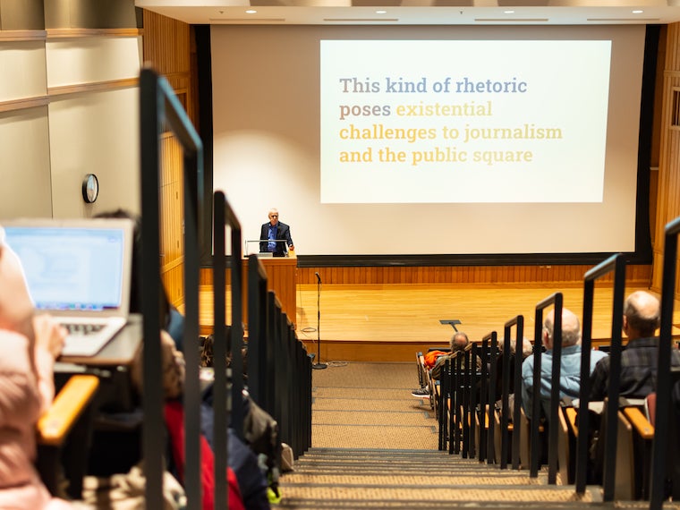 A man stands at a podium with large slides on a screen behind him.