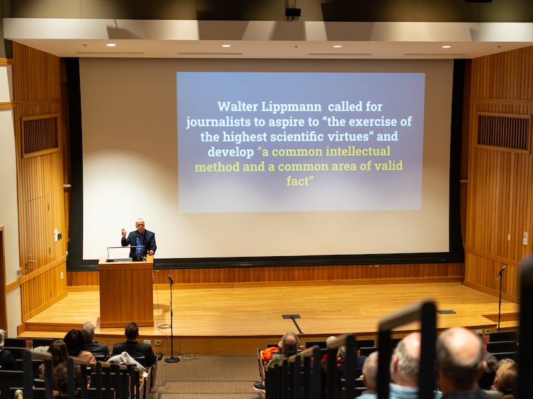 A man stands at a podium with large slides on a screen behind him.