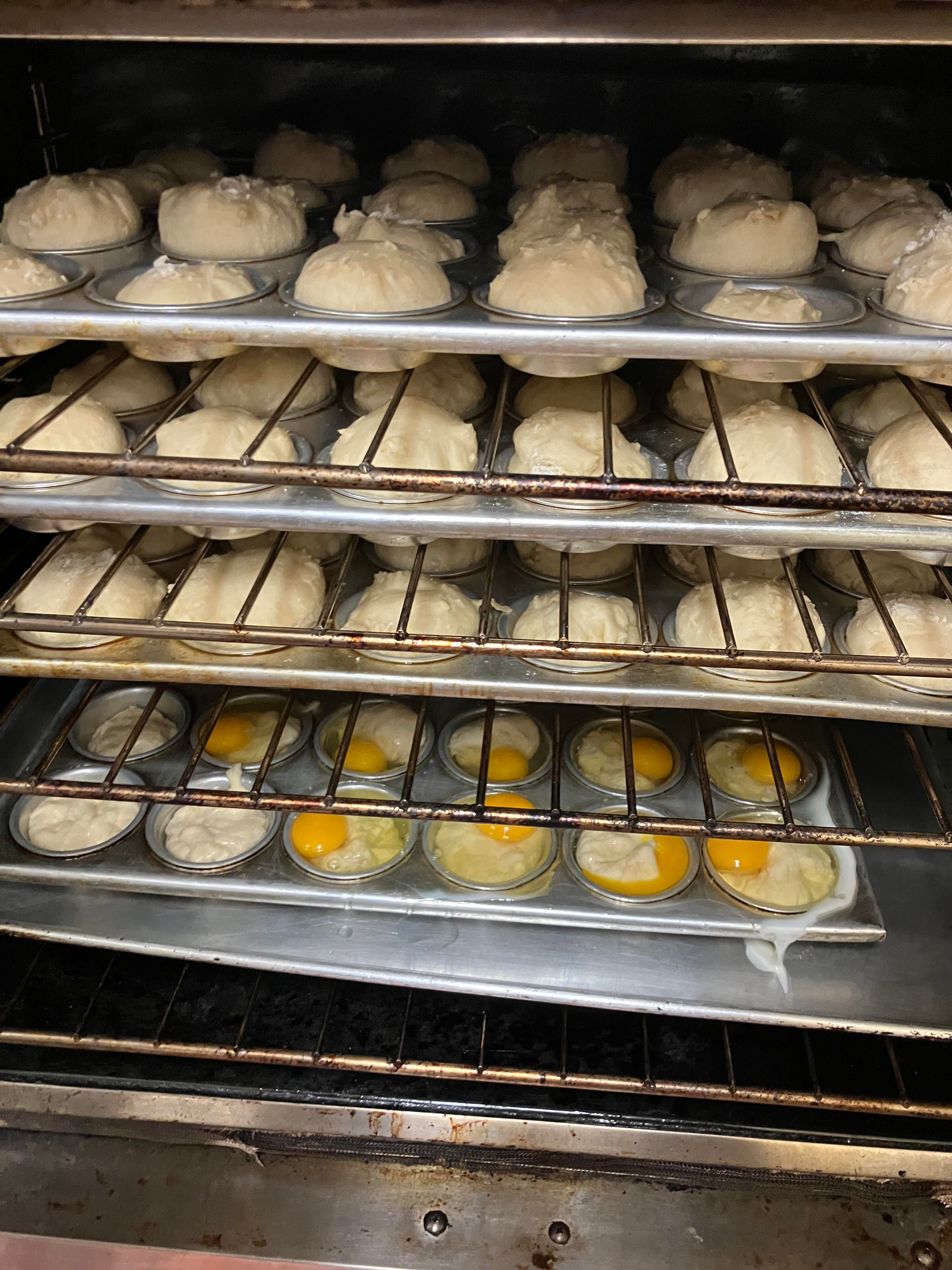 Picture of the inside of an oven. There are four racks. On the upper three racks there are rolls baking in muffin tins. On the lowest rack there are raw sunny side up eggs baking in muffin tins.