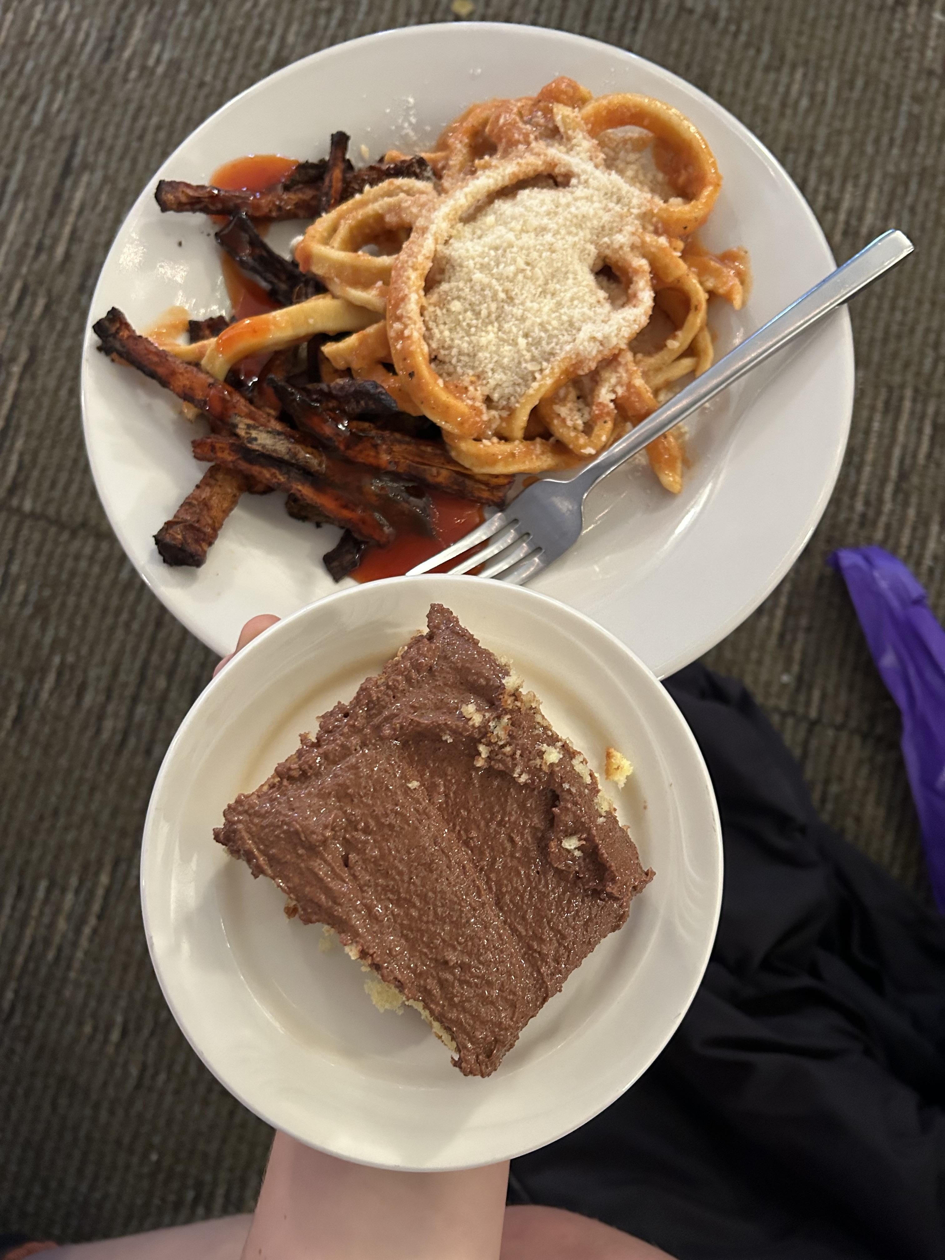 Two round white plates are pictured. On one is handmade pasta with parmesan and cooked carrot sticks. On another is a square piece of chocolate frosted cake.