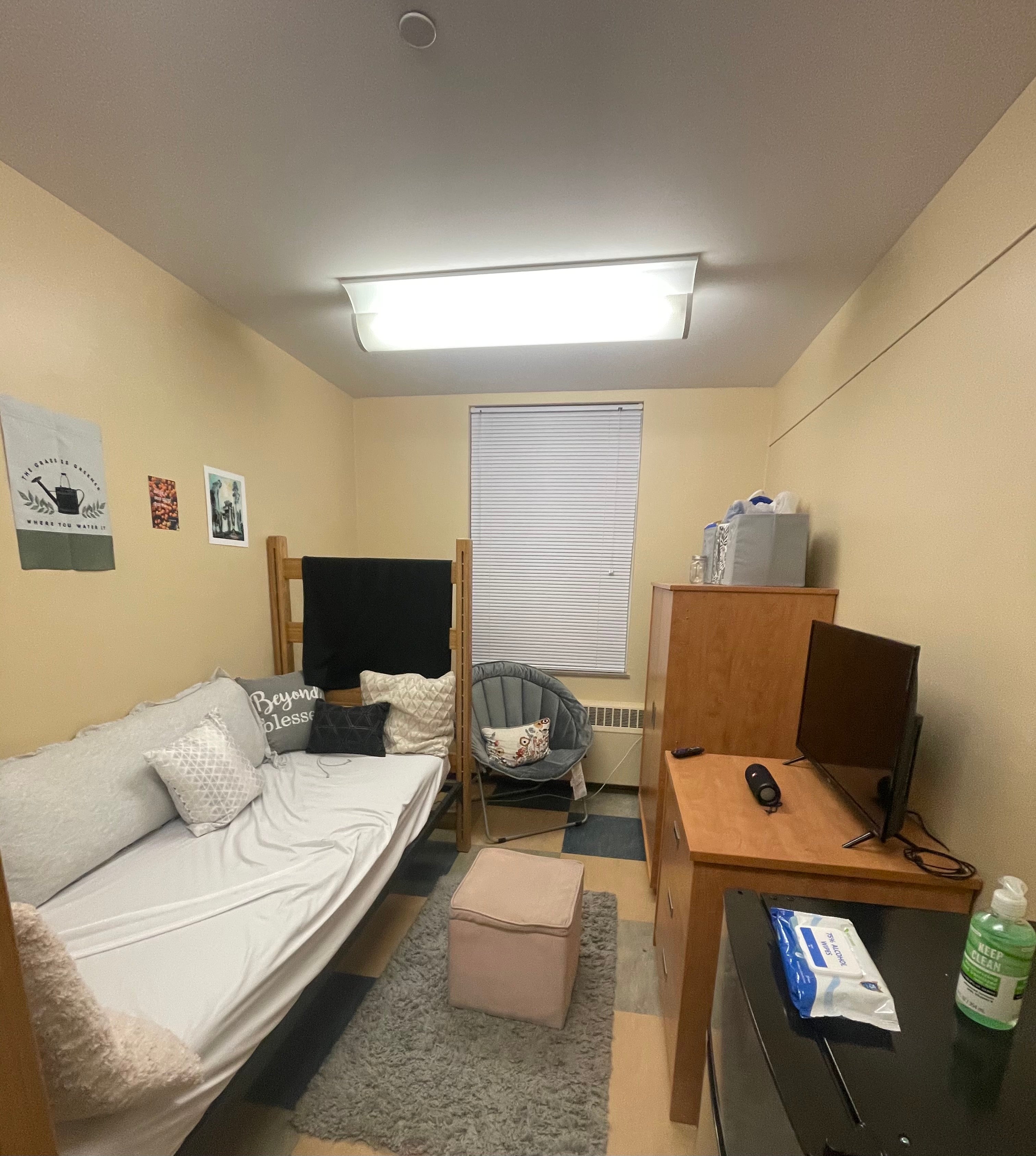 This pictures shows the other half of a decorated divided double. The dorm bed is pushed up against the left wall with many pillows lining the wall and the bed frame. The bed is made to look like a couch with wall art above it. A light pink ottoman sits on a gray rug in front of the bed. A gray chair is placed in the far left corner across from a wardrobe. A television rests on top of a smaller cabinet opposite the dorm bed. A mini fridge is in the foreground of the image, located against the ri