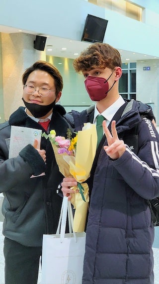 My friend and I in the lobby of the concert hall standing together after our concert. I am holding flowers and a gift bag and he is holding a letter.