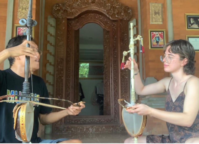 Ilana and an old man sit on the ground facing each other and holding string instruments with bows outside of a house