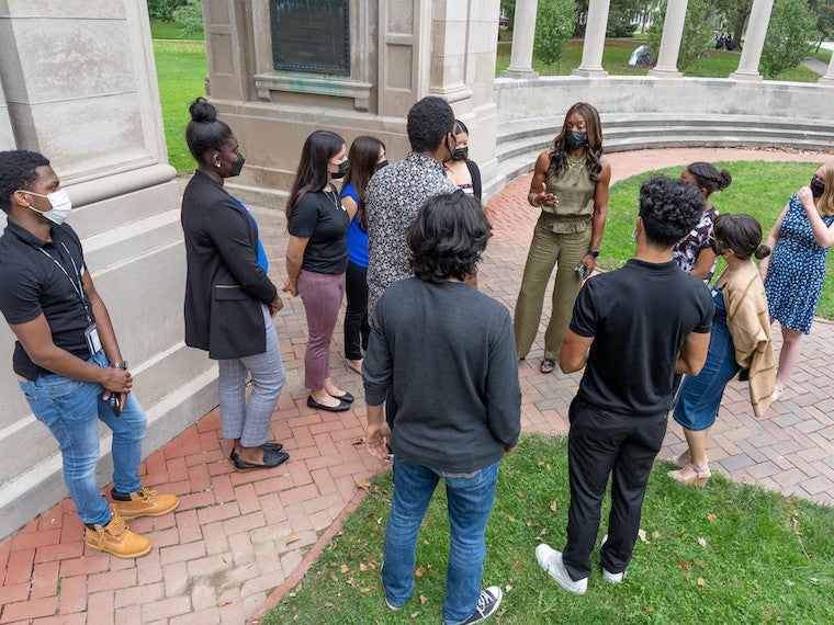 A group of 10 students listen to a woman speaking to them.