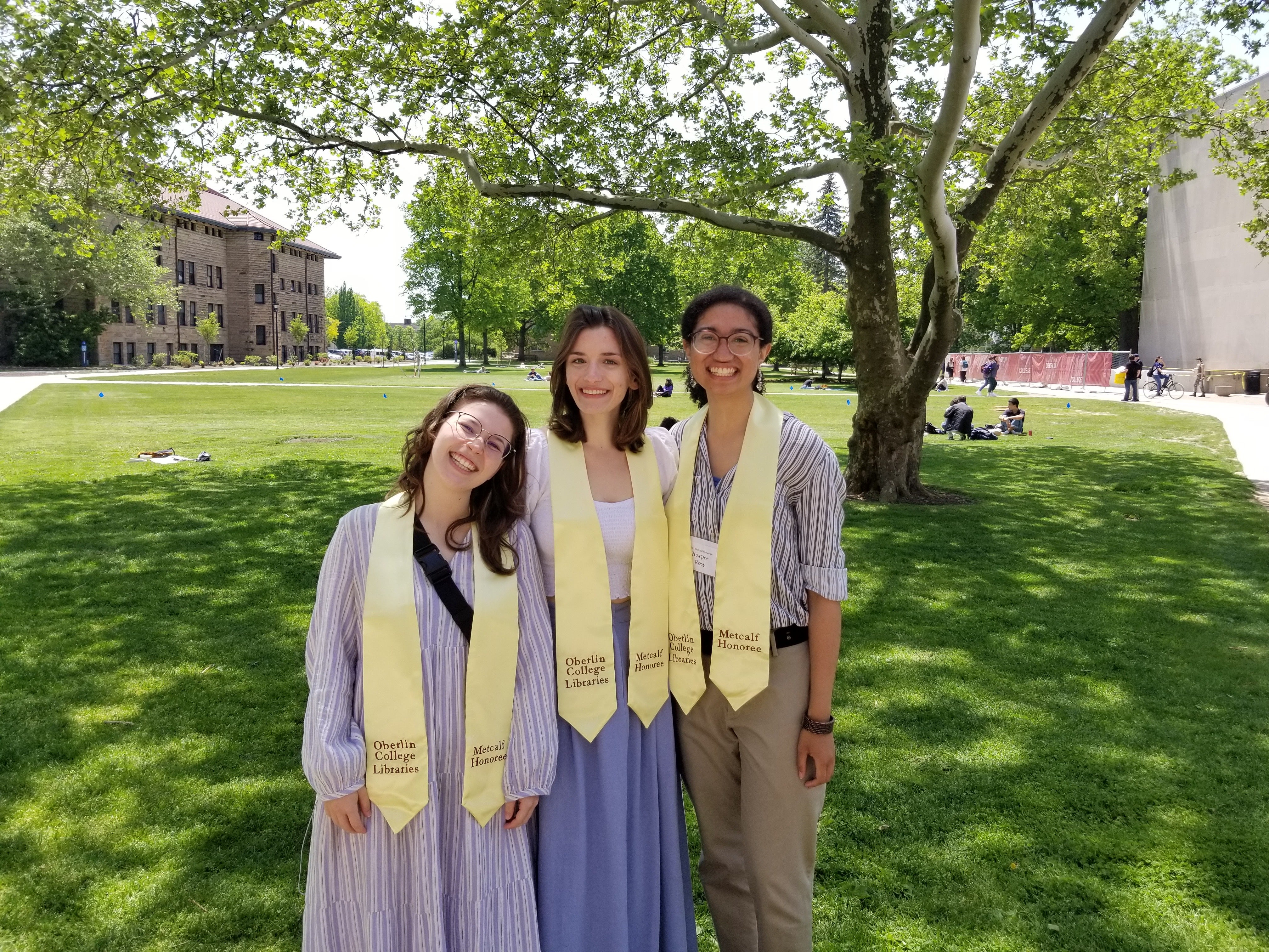 Megan stands between two other people in the middle of Wilder Bowl on a sunny day. All three individuals are wearing gold sashes around their shoulders that read "Oberlin College Libraries" on one side and "Metcalf Honoree" on the other. They are smiling and having a great time!!