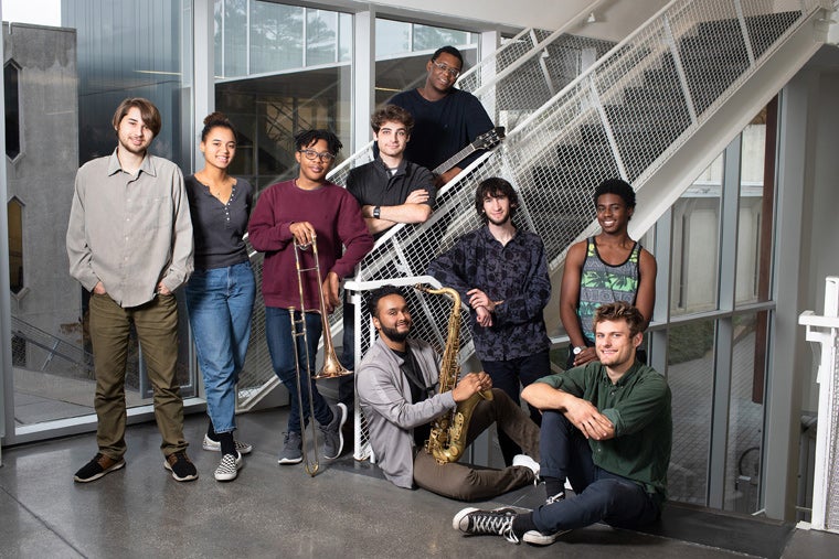 Group photo of the 9 ensemble members, three of them holding instruments: a tenor sax, a trombone, and an electric guitar.