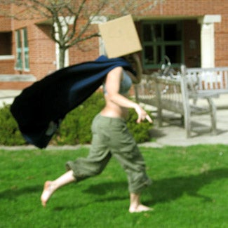 Her identity concealed by a cardboard box over her head, a woman runs in the grass, her cape flapping behind her.