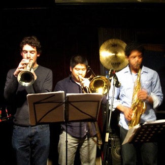 Three musicians play behind a music stand - trumpet, trombone, and alto sax